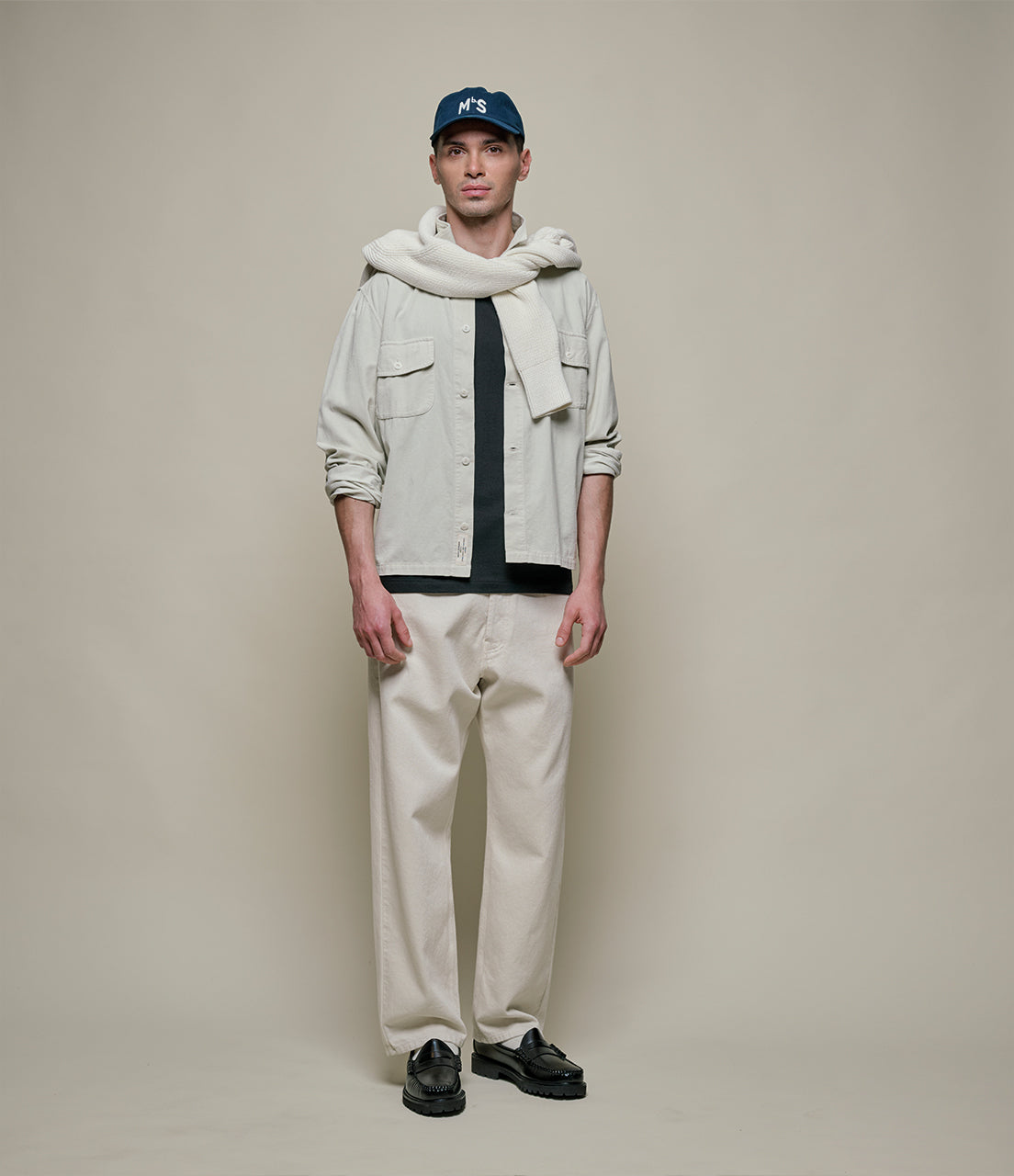 Man wearing a light jacket, scarf, cap, black shirt, beige pants, and black shoes on a plain background