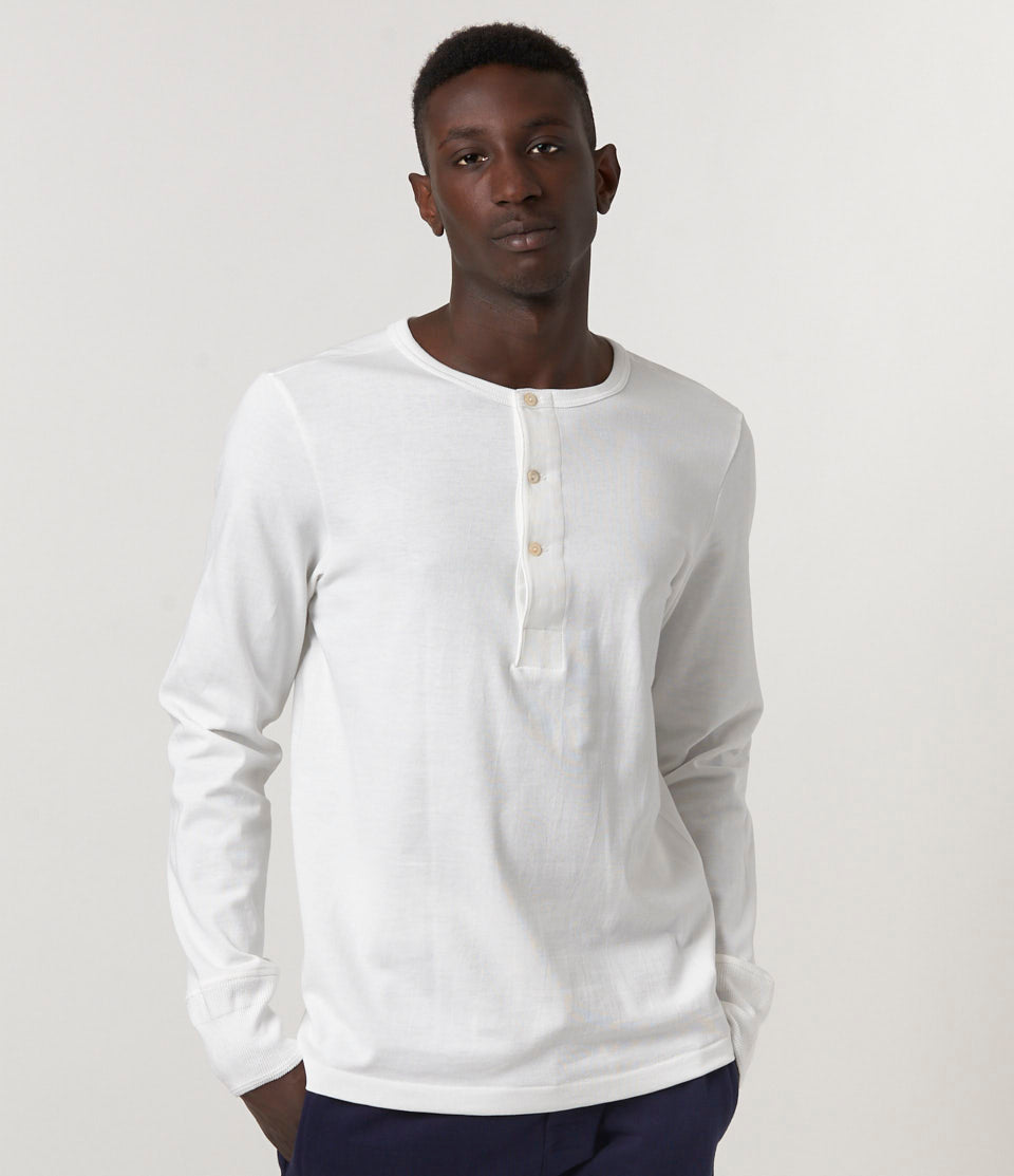 A male model wearing a white long sleeve henley shirt with buttons at the neck and a relaxed fit.