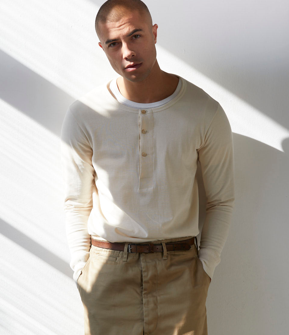 A male model wearing a beige long sleeve henley shirt with buttoned neckline and a pair of beige trousers.