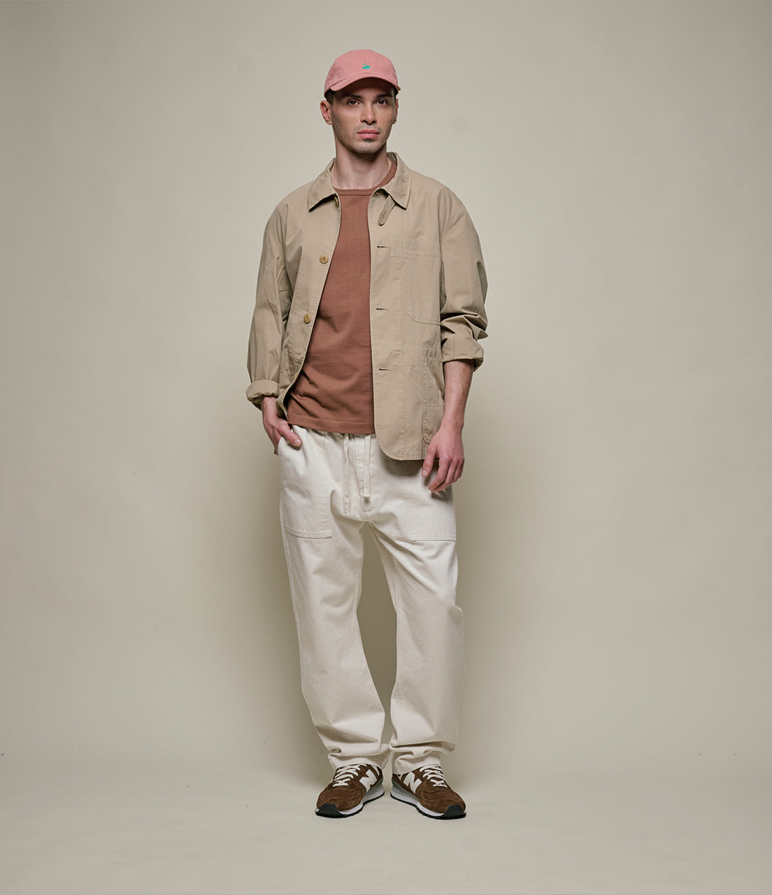 Man wearing a beige jacket, brown shirt, white pants, and pink cap on a plain background