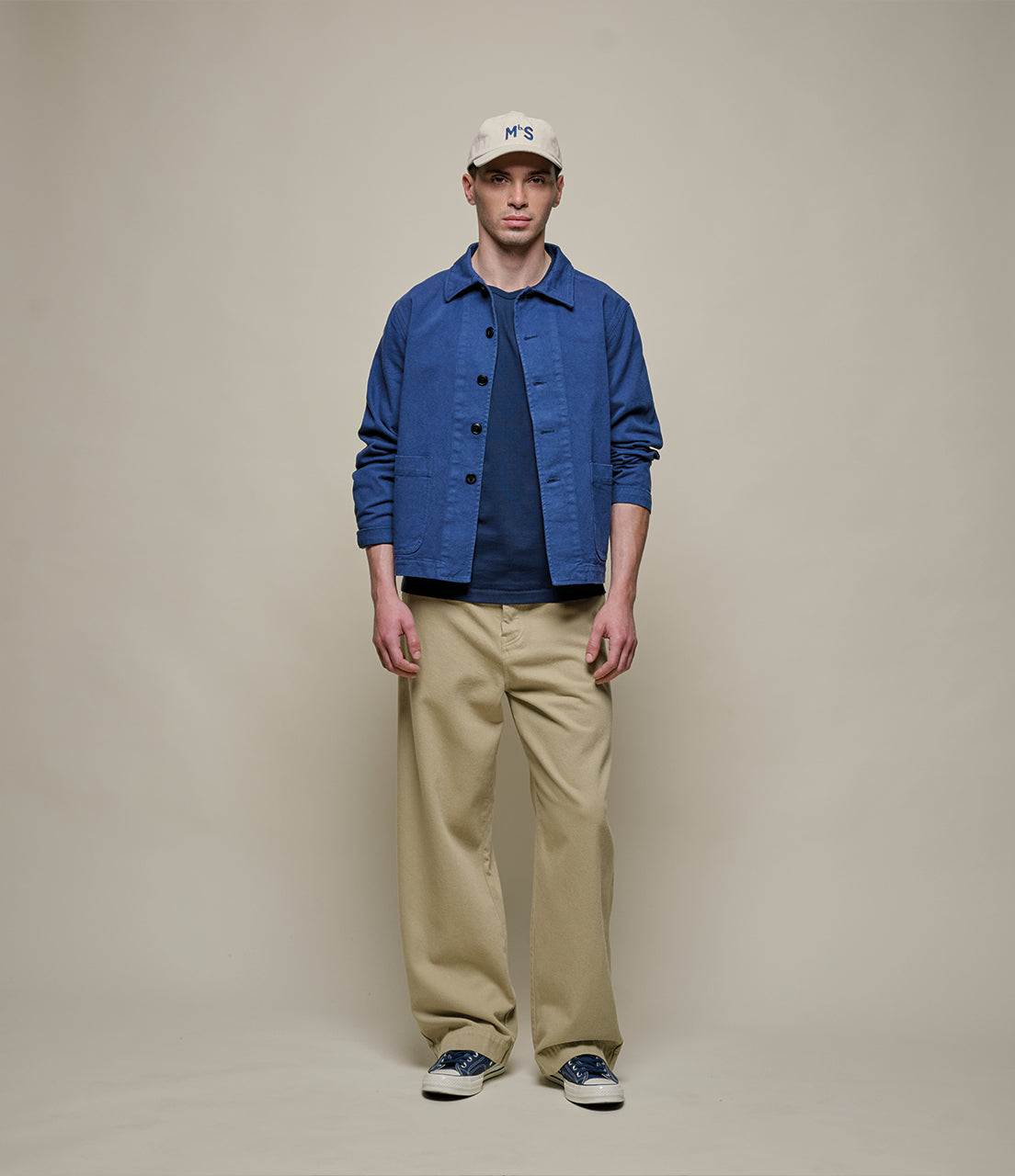 Man wearing a blue jacket, navy shirt, beige pants, and white cap against a plain background