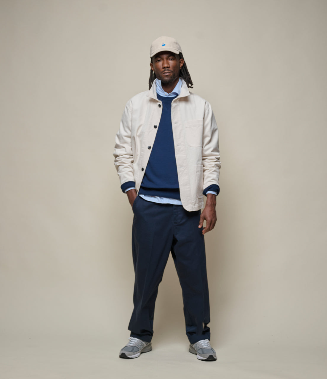 Man wearing a white jacket, blue sweater, and navy pants against a beige background