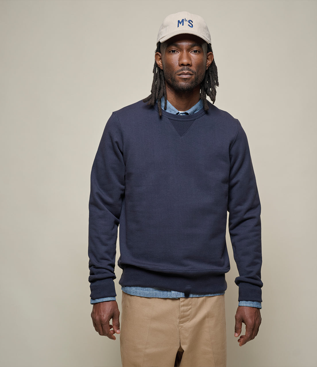 Man wearing a navy sweater, beige pants, and a white cap with 'MbS' logo on a plain background