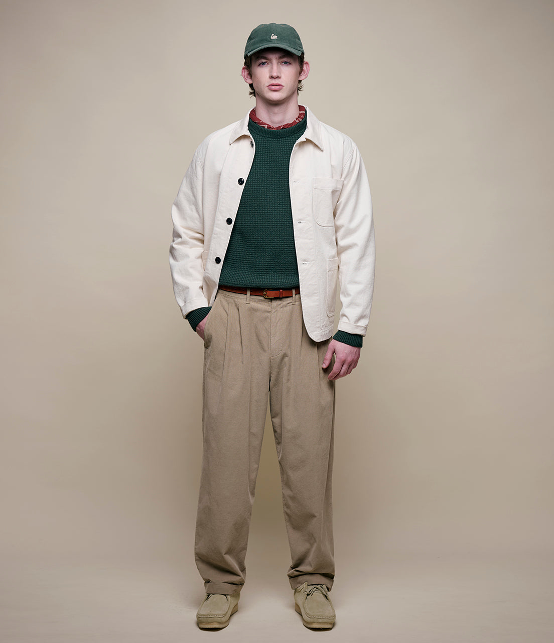 Man wearing a cream jacket, green cap, dark shirt, beige pants, and brown shoes on a beige background