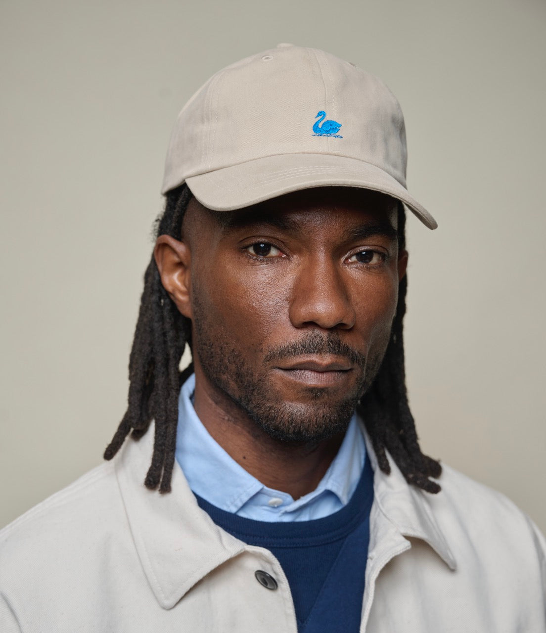 Man wearing a beige cap with a logo, light jacket, and blue shirt against a plain background