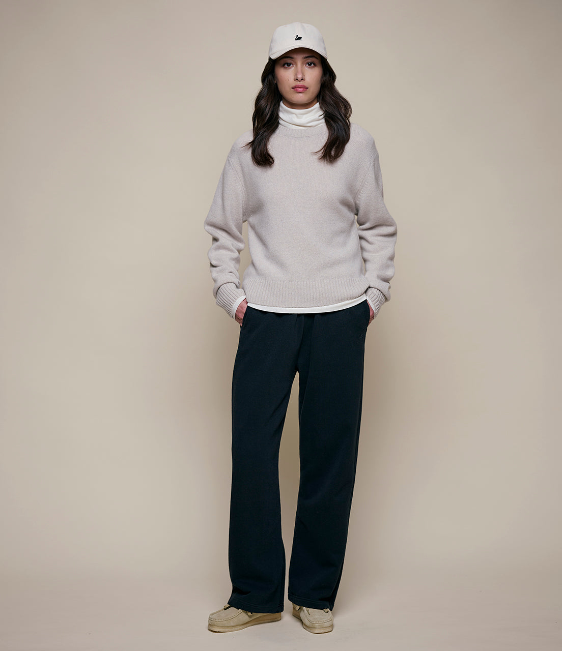 Person wearing a light gray sweater, black pants, and a white cap on a beige background