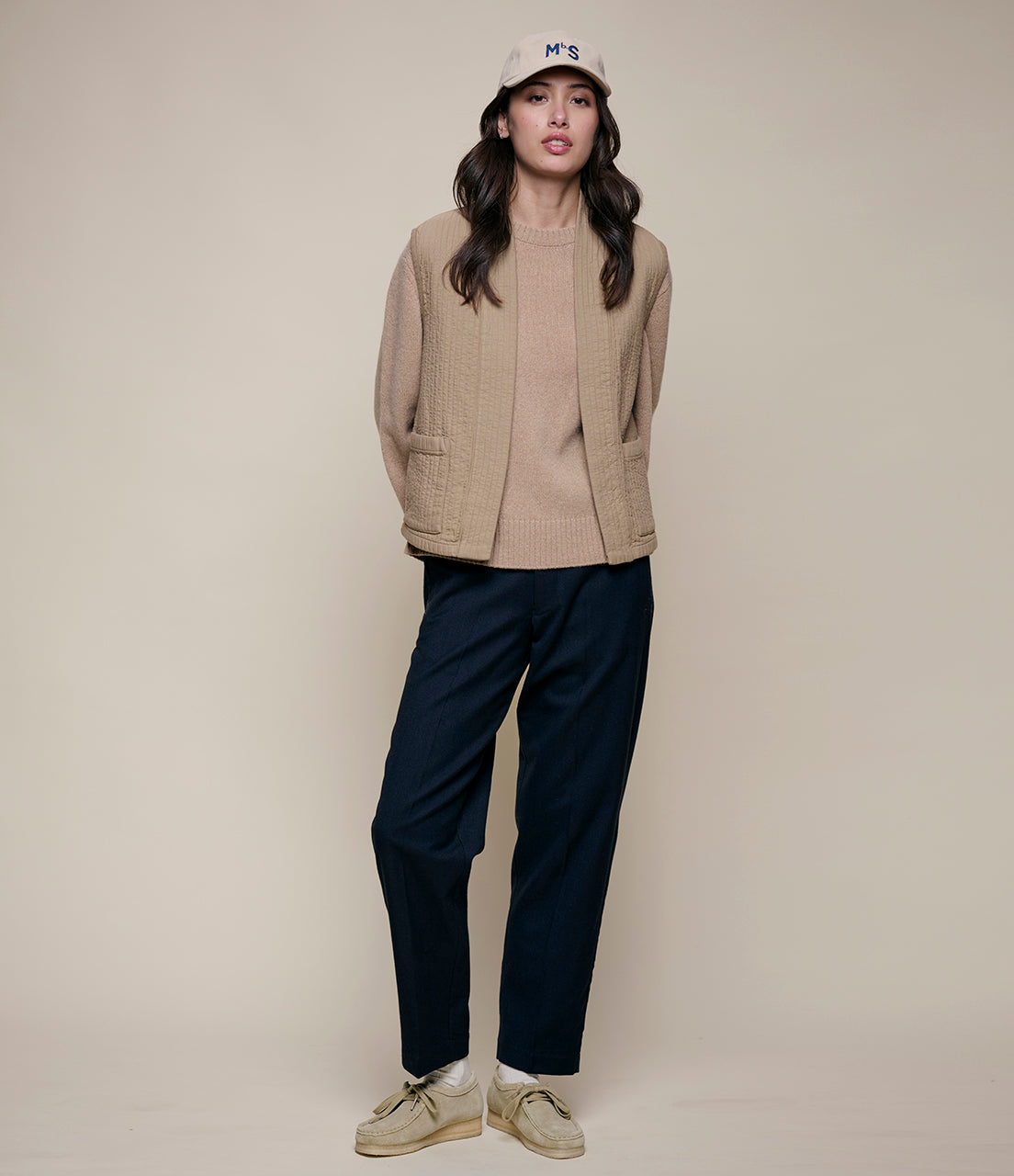 Person wearing a beige vest, beige sweater, navy pants, and white sneakers on a plain background