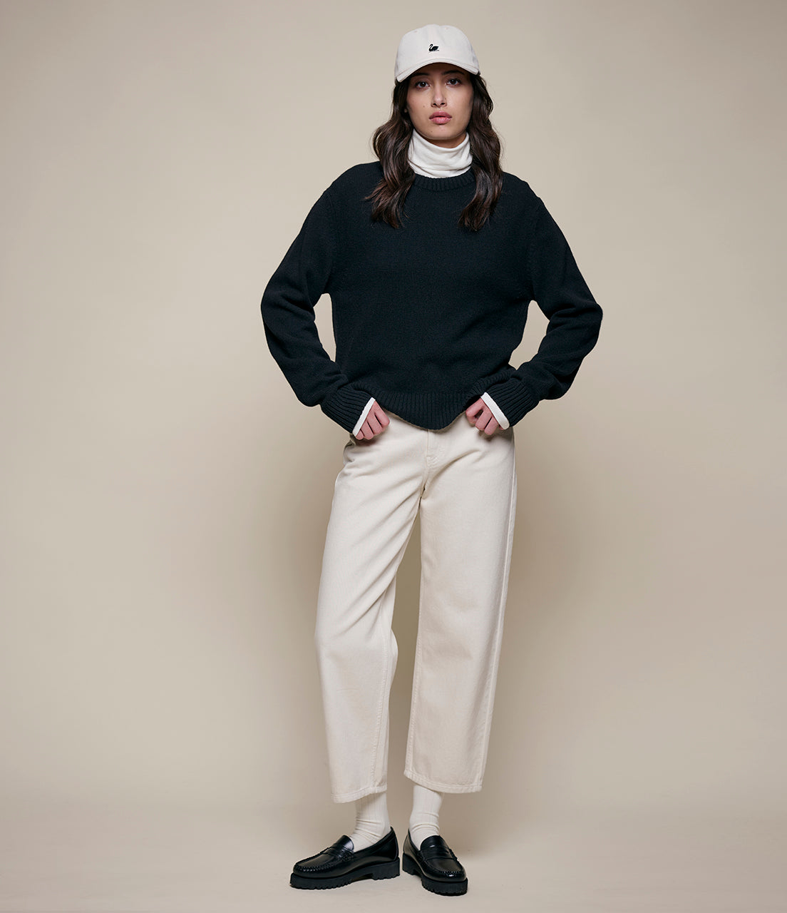 Person wearing a black sweater, white pants, and a white cap on a beige background