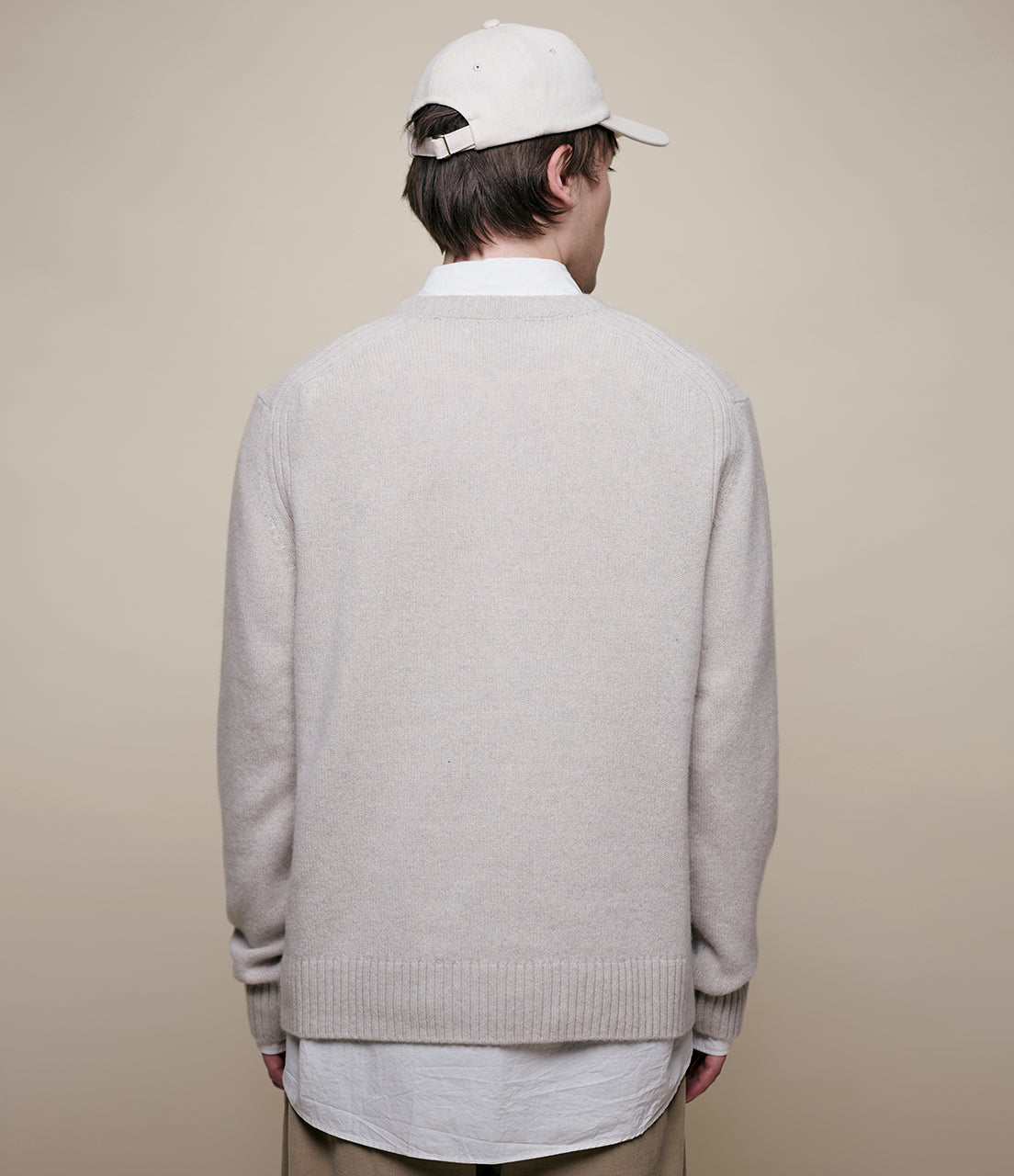 Person wearing a light gray sweater and white cap from behind on a beige background