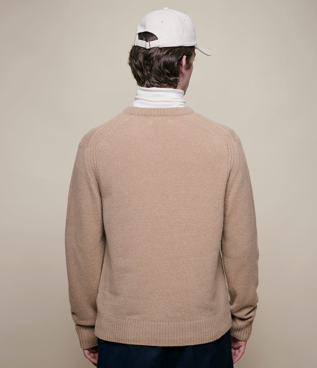 Person wearing a beige sweater and white cap from behind on a beige background