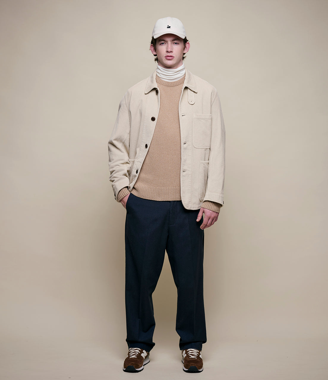 Man wearing a beige jacket, brown sweater, and dark pants against a plain background