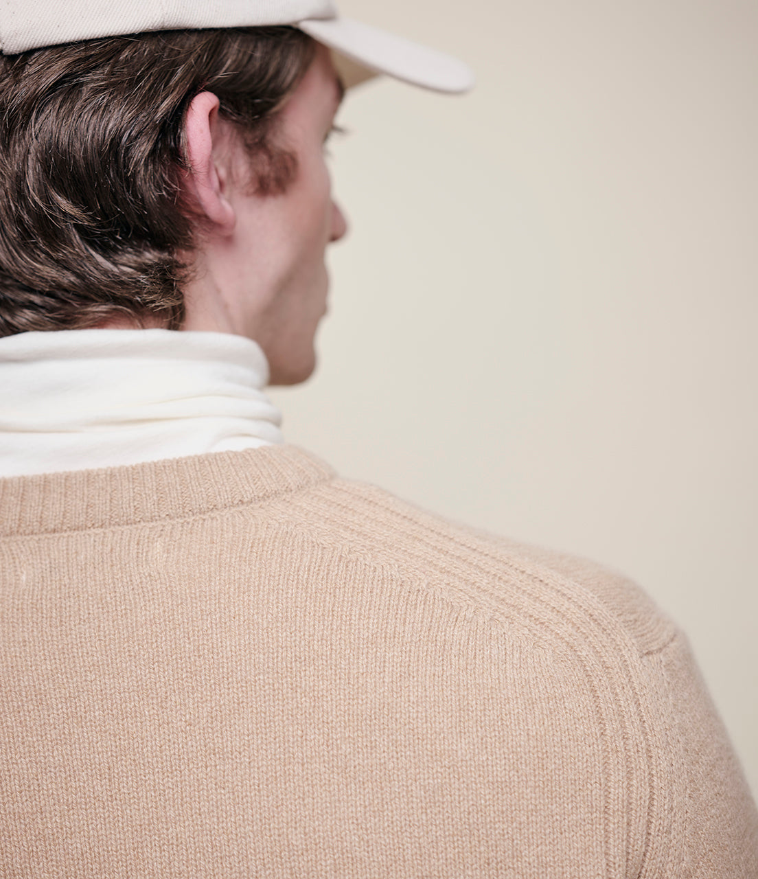 Person wearing a beige cap and light brown sweater with a white turtleneck underneath, standing against a plain background.
