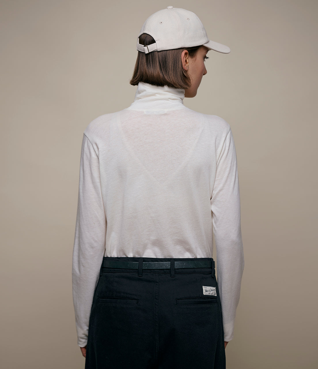 Person wearing a white turtleneck, black pants, and a white cap on a beige background