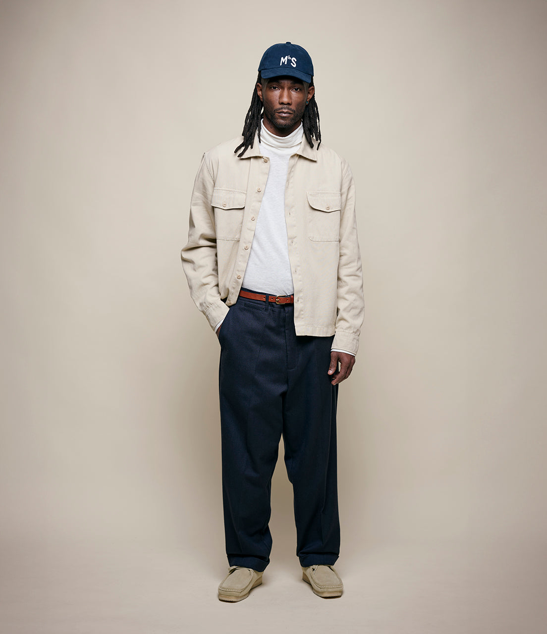 Person wearing a beige jacket, navy pants, and a blue cap on a plain background
