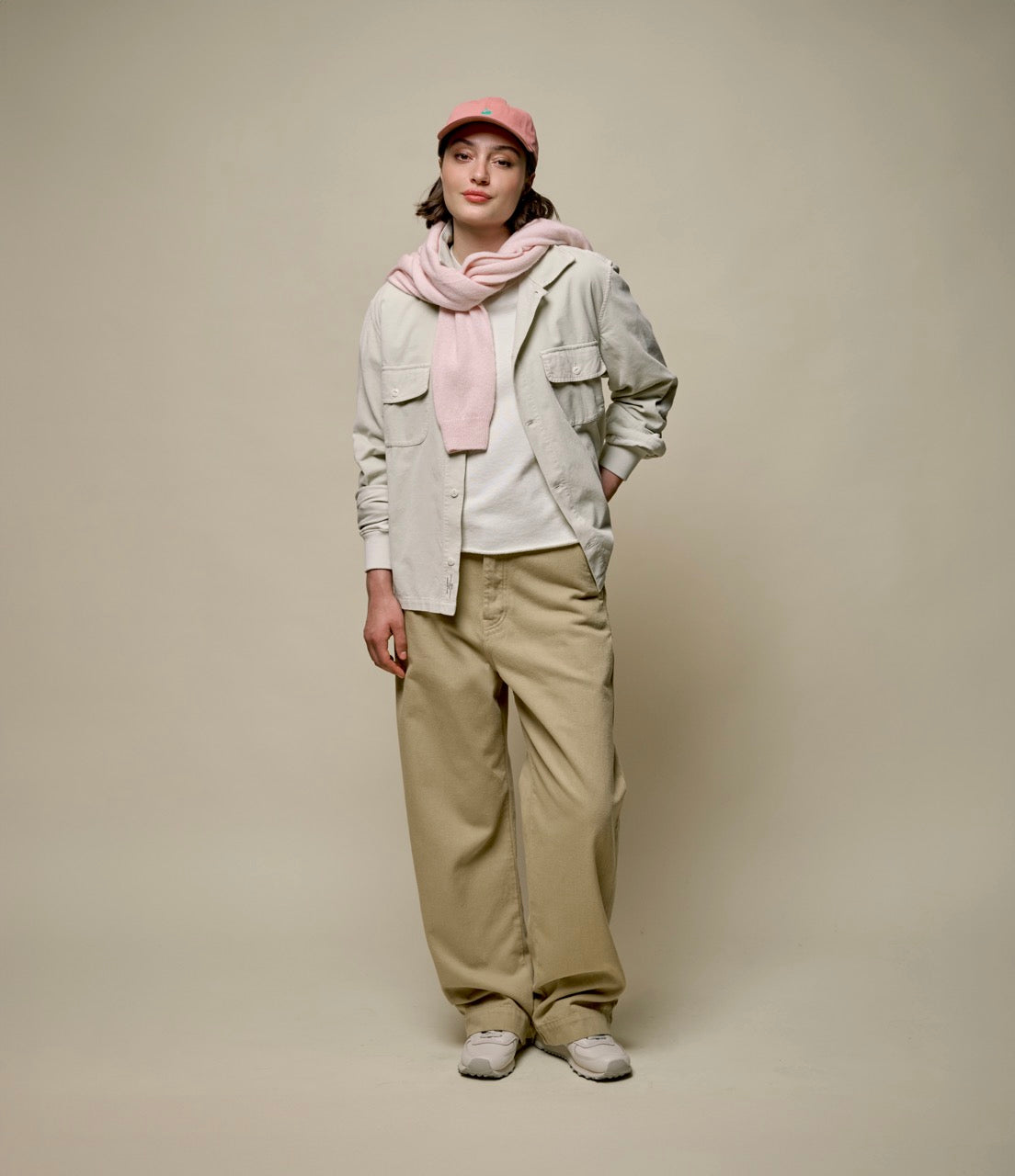 Person wearing a light gray jacket, beige pants, pink scarf, and cap against a plain background.