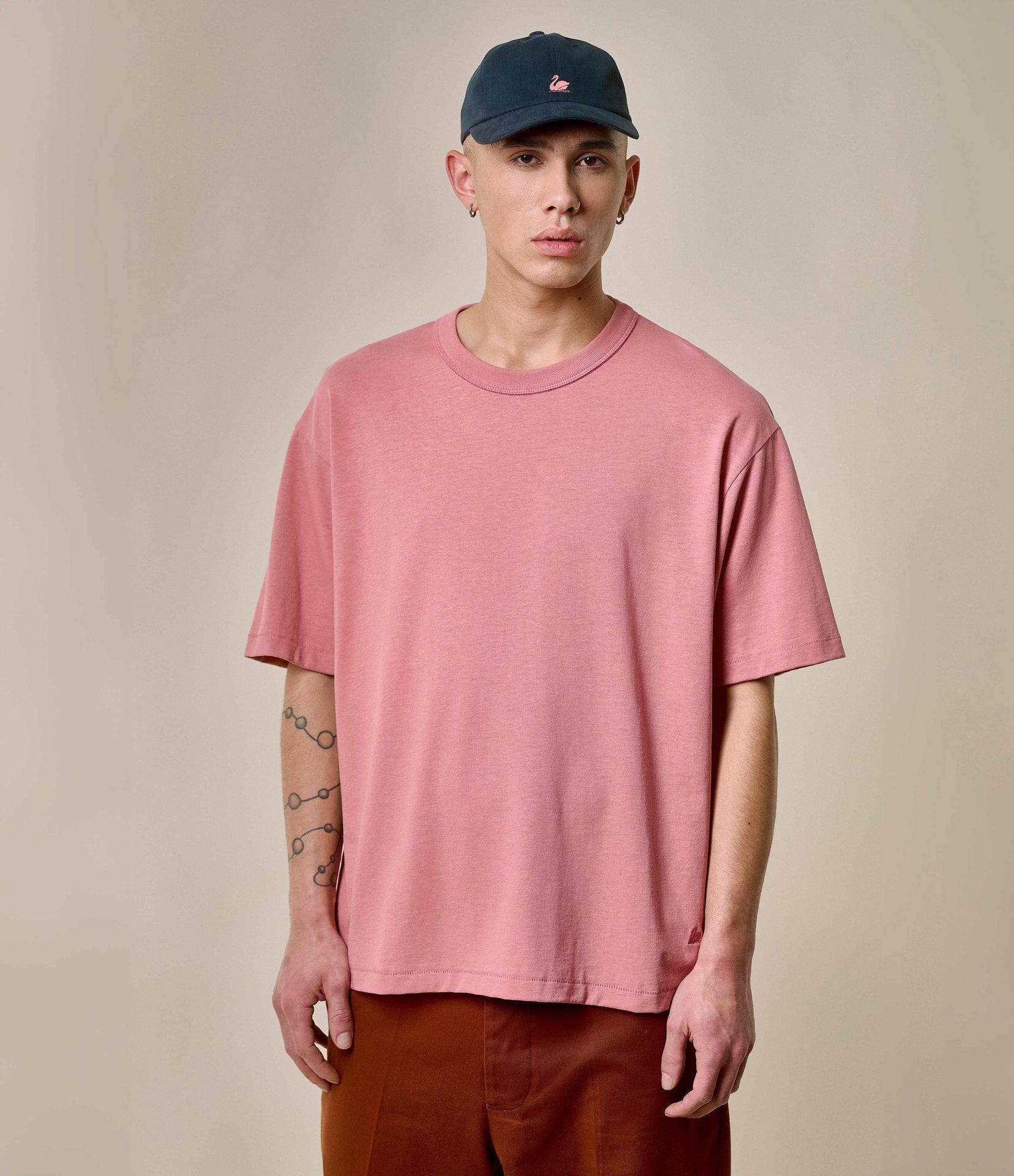 A person wearing a pink oversized crew-neck T-shirt with short sleeves, paired with brown trousers and a blue cap.