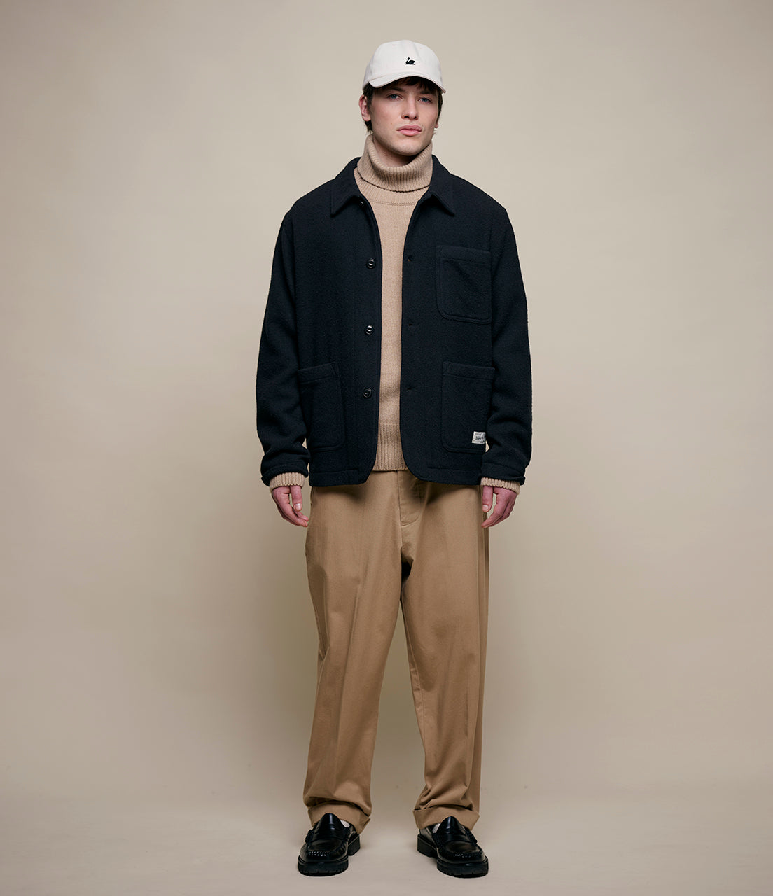 Man wearing a black jacket, beige sweater, and pants with a white cap on a beige background