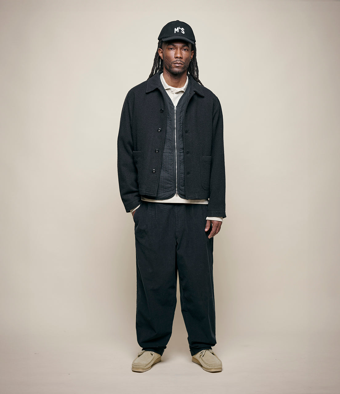 Man wearing a dark jacket and pants with a cap on a plain background