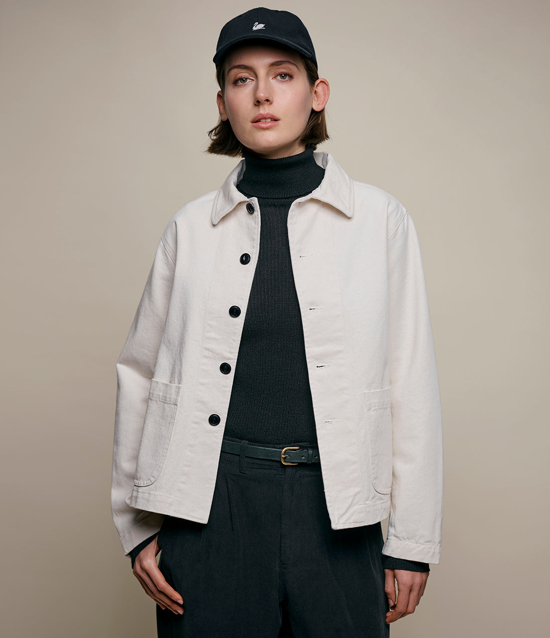 A person wearing a cropped twill jacket in a light color, with a pointed collar, buttoned front, and a waist belt.
