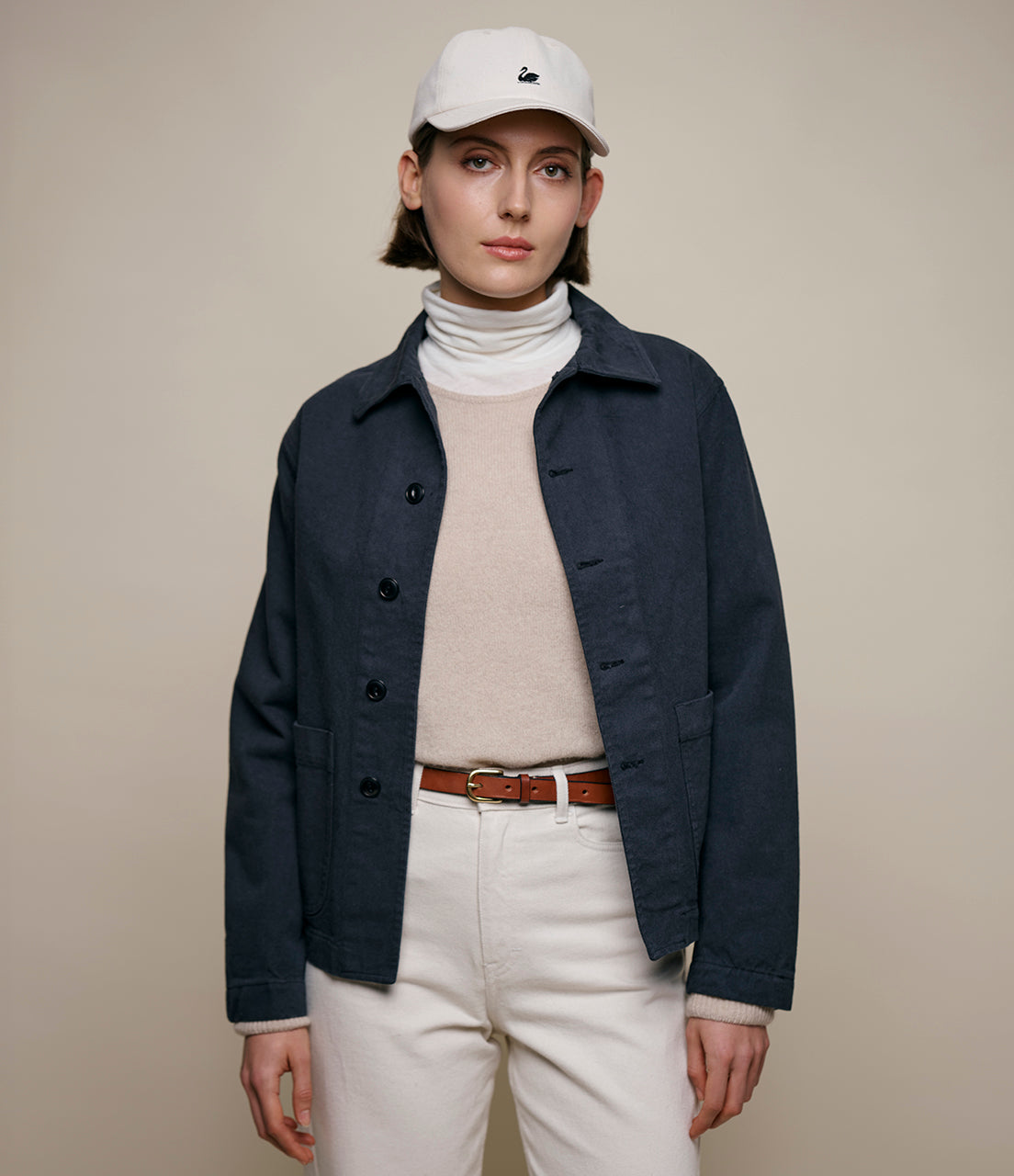 A person wearing a dark grey cropped twill jacket with a collar, buttoned front, and light-colored trousers. They are also wearing a white cap and a light pink turtleneck.
