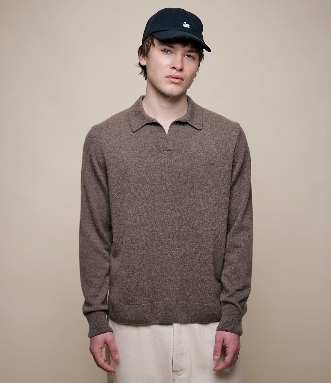 A male model wearing a long sleeve polo shirt in a beige color with a V-neck collar. The model is also wearing a black cap and light-colored trousers.
