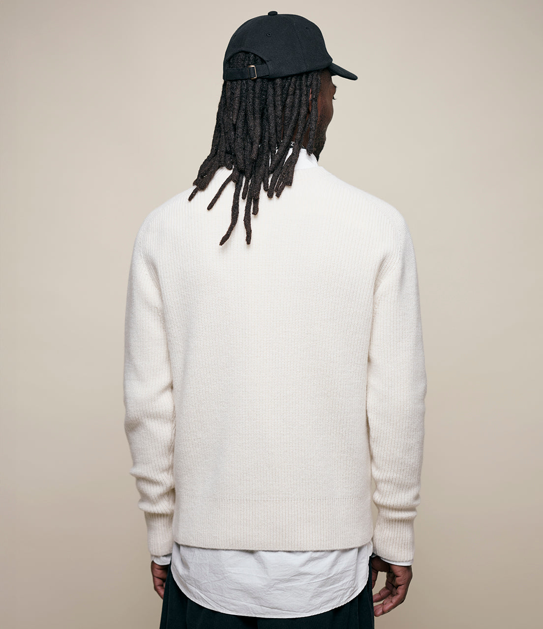 Person wearing a white sweater and black cap from behind on a beige background