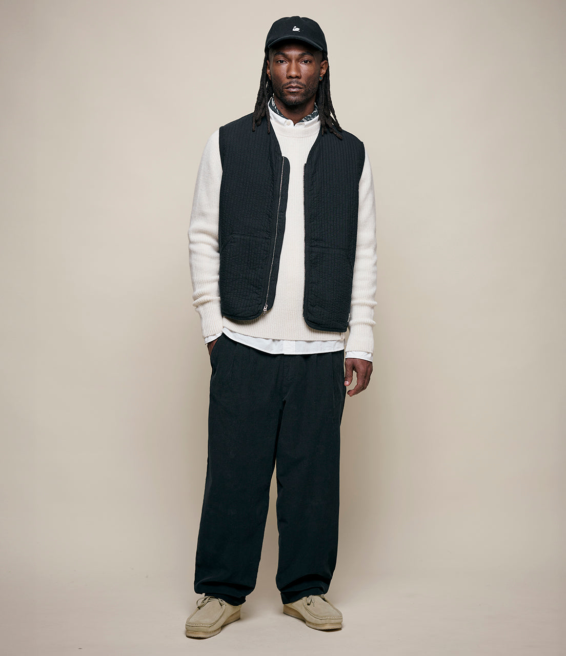 Man wearing a black vest over a white sweater with dark pants on a beige background