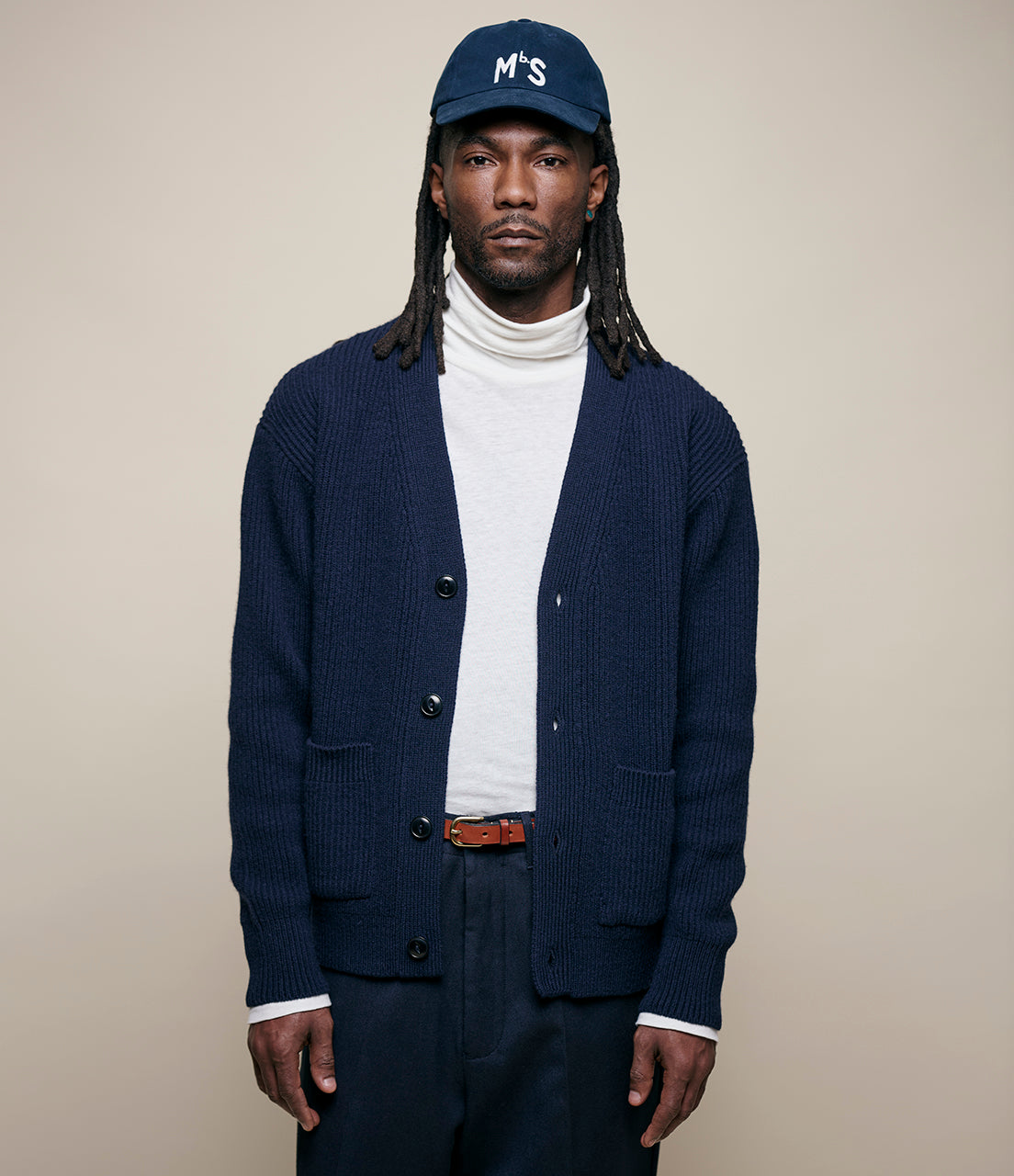 Man wearing a navy cardigan, white turtleneck, and dark pants against a beige background