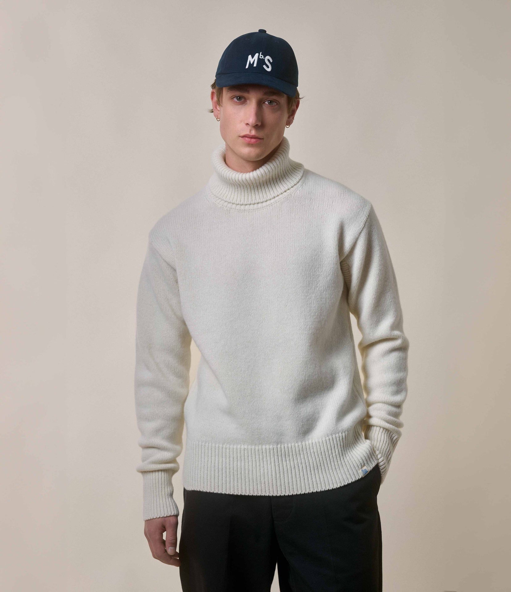 Person wearing a white turtleneck sweater and black cap on a plain background