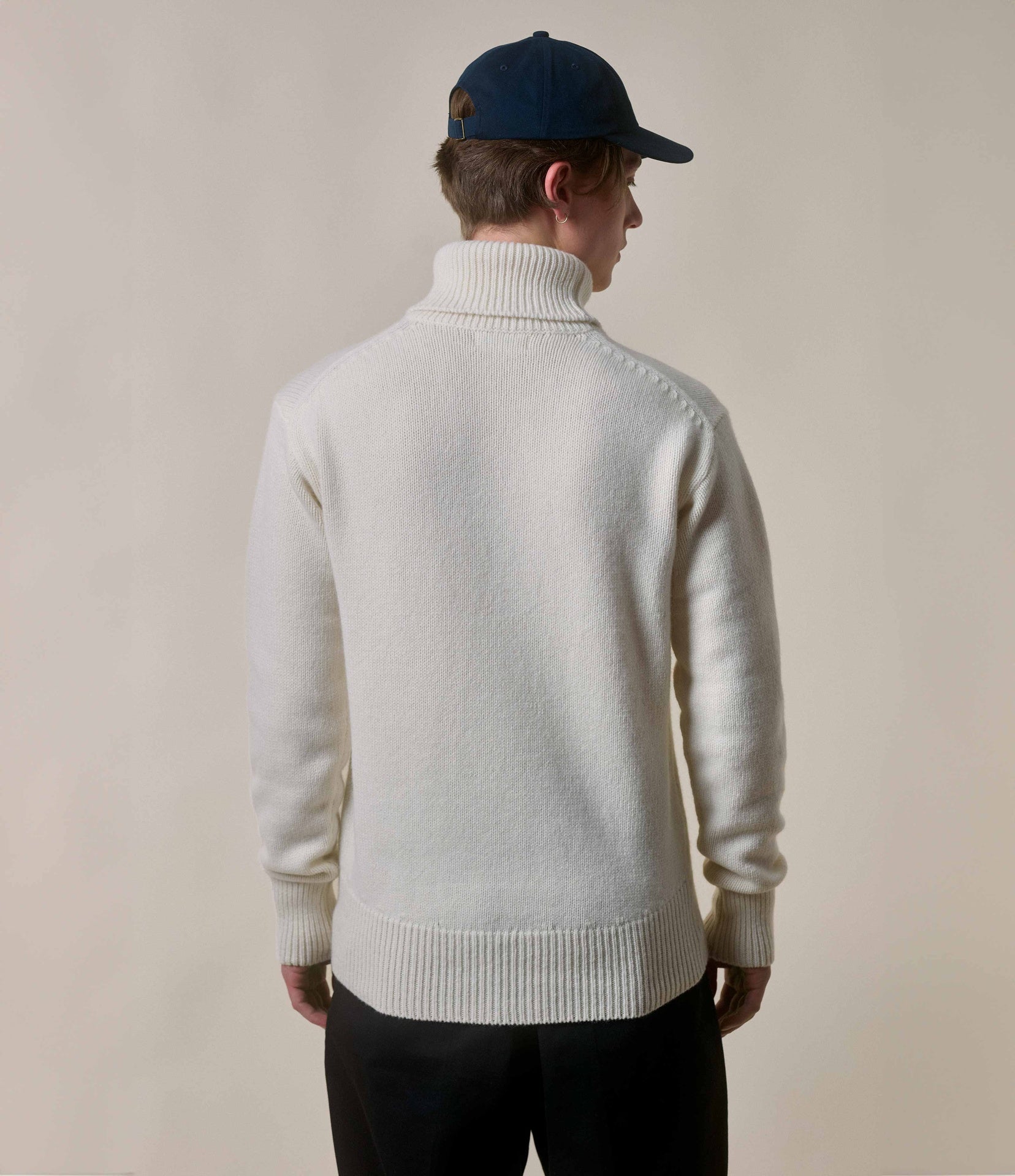 Person wearing a white turtleneck sweater and dark cap on a plain background