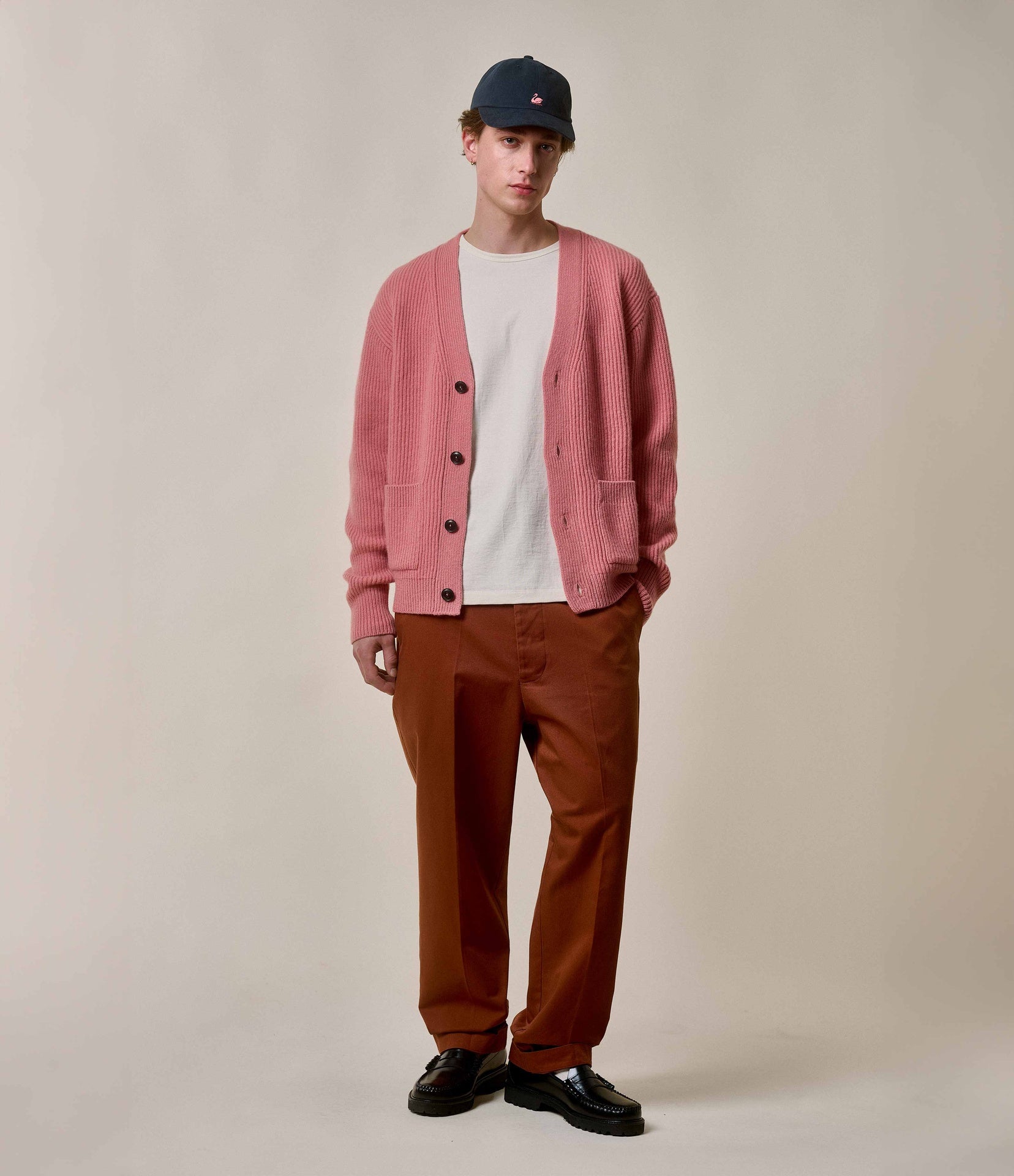 Person wearing a pink cardigan, white shirt, brown pants, and black shoes on a beige background