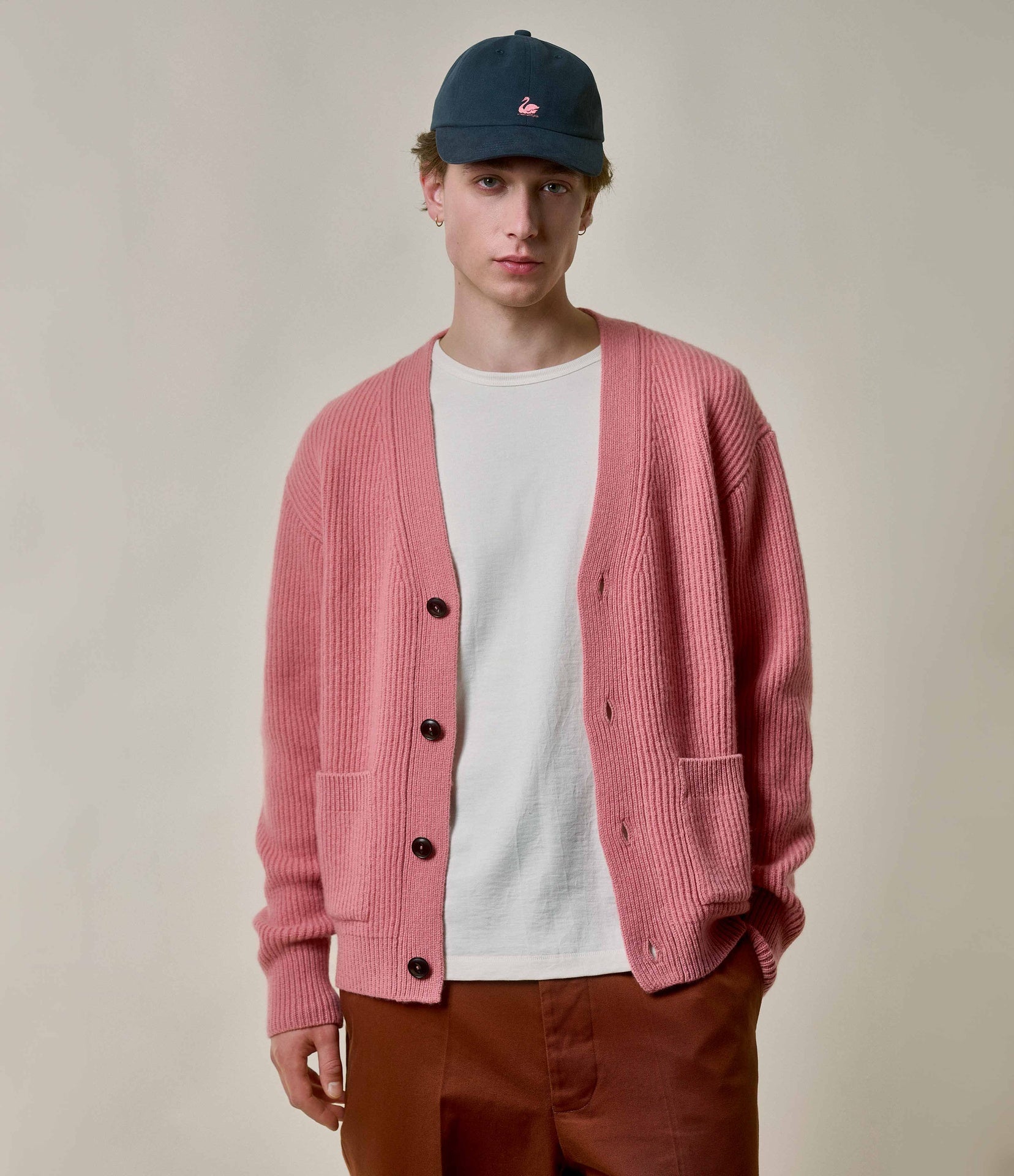 Person wearing a pink cardigan, white t-shirt, and brown pants with a blue cap on a plain background