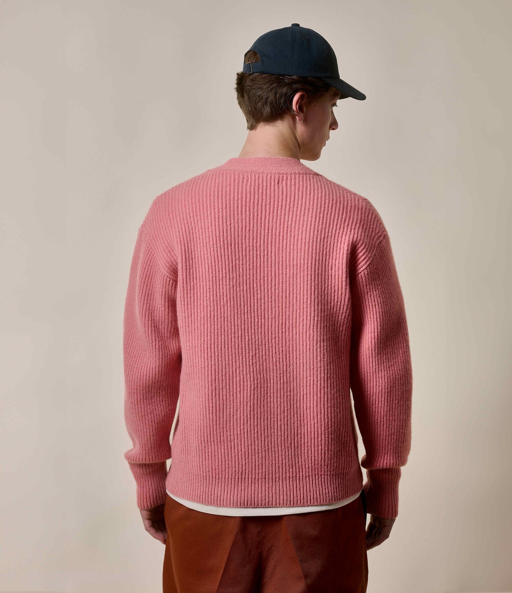 Person wearing a pink cardigan and brown pants with a blue cap on a plain background