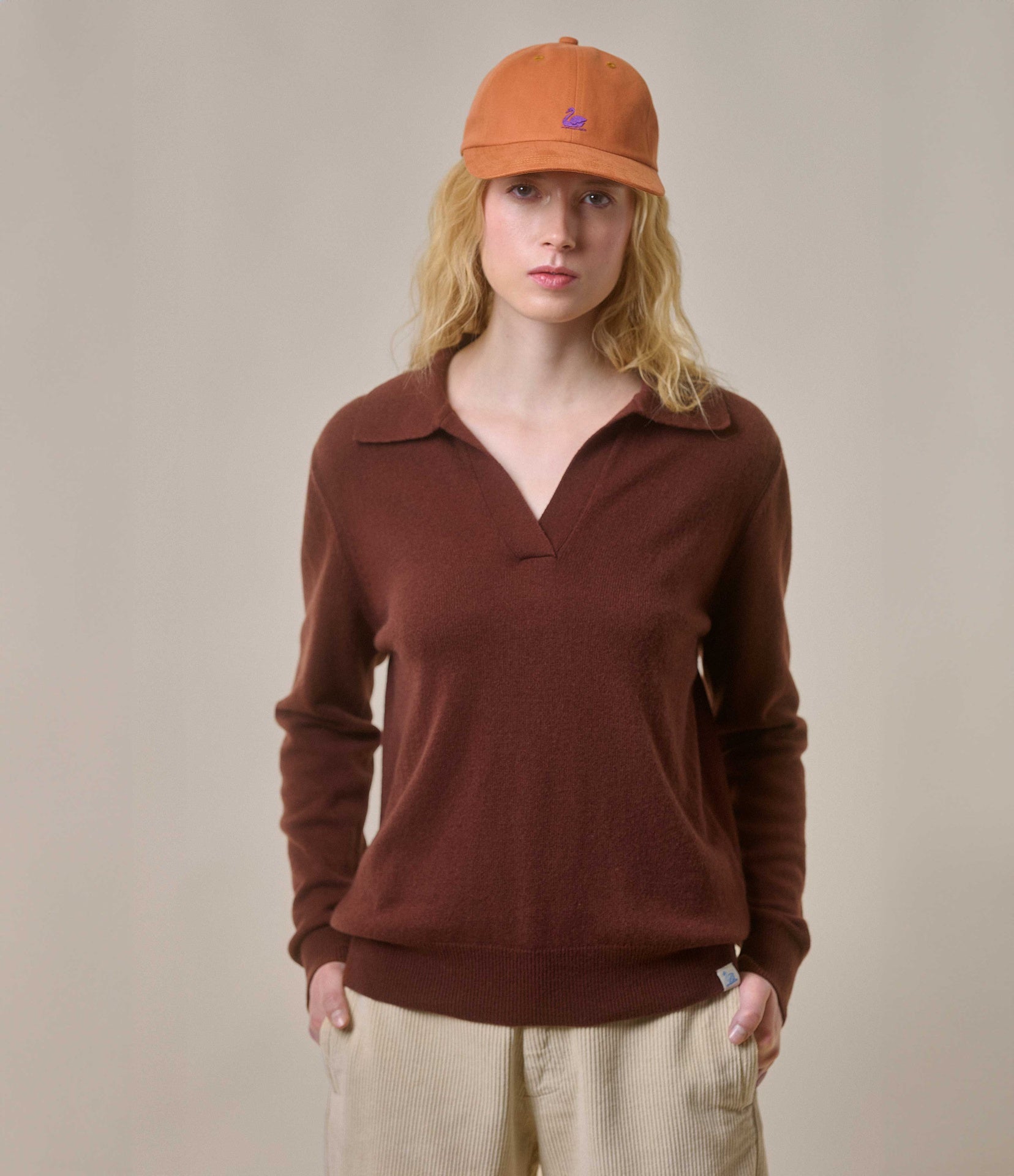 A person wearing a brown V-neck sweater with long sleeves, paired with light-colored pants and an orange cap.