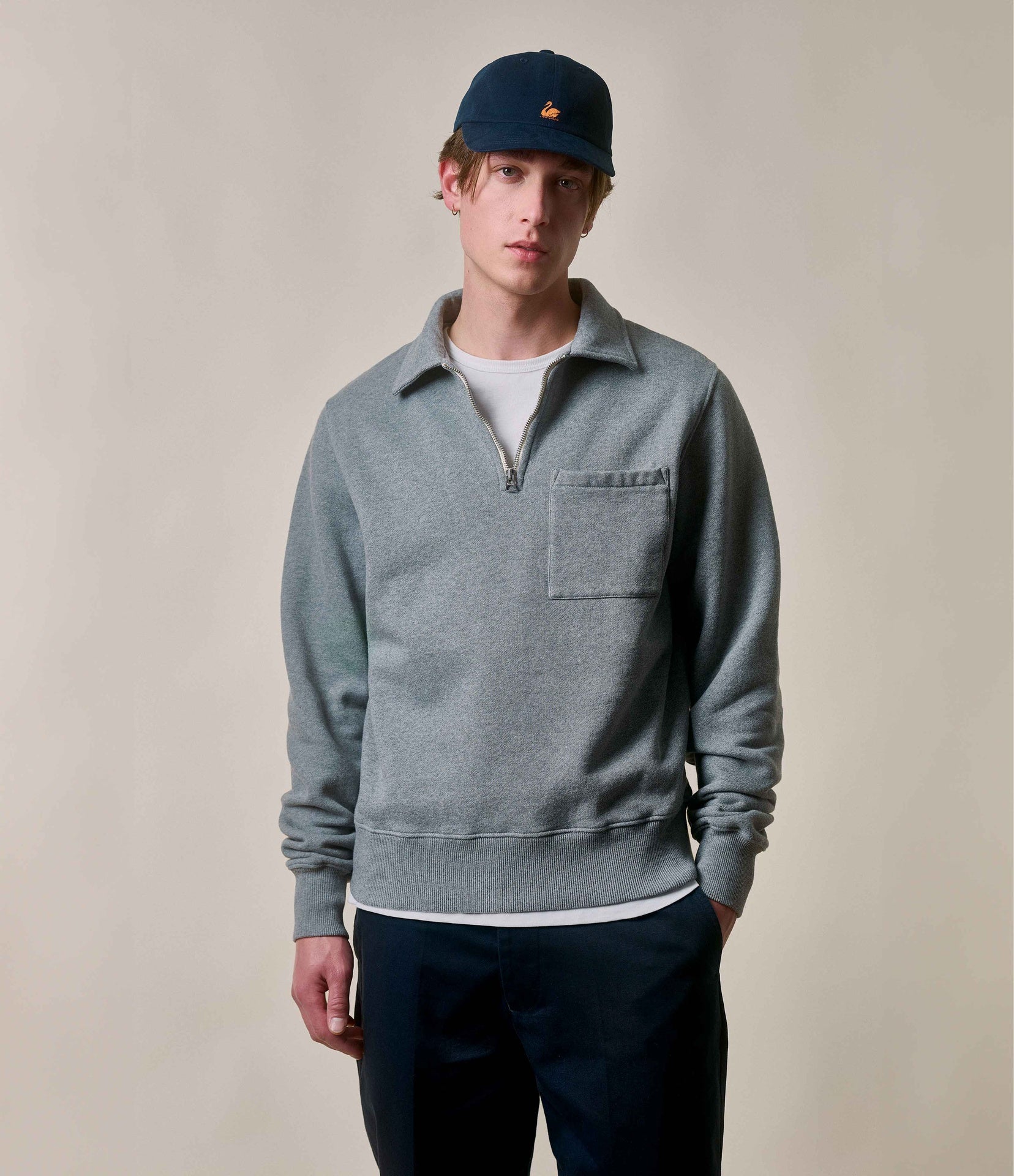 A male model wearing a gray vintage fleece half-zip sweatshirt with a polo collar and a chest pocket, paired with dark pants and a blue cap.