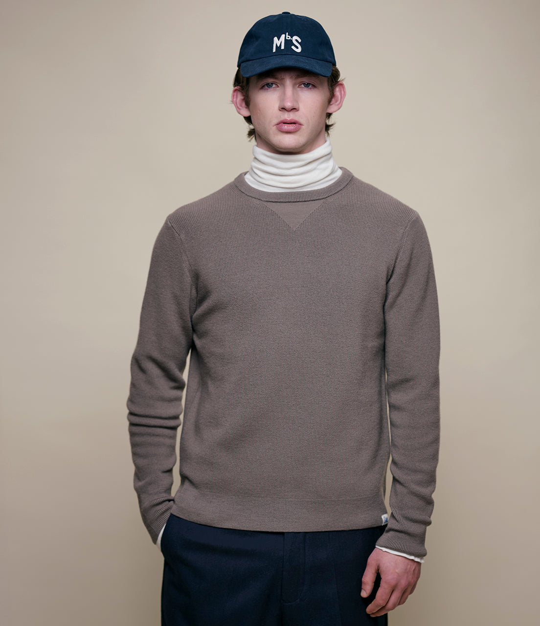 Person wearing a gray sweater, white turtleneck, and navy cap on a beige background