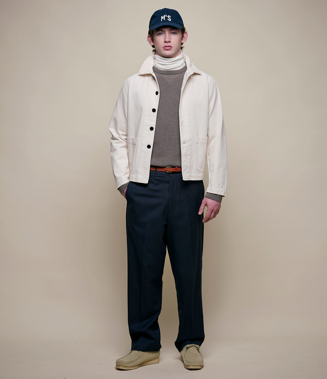 Person wearing a white jacket, gray sweater, dark pants, and a cap on a beige background
