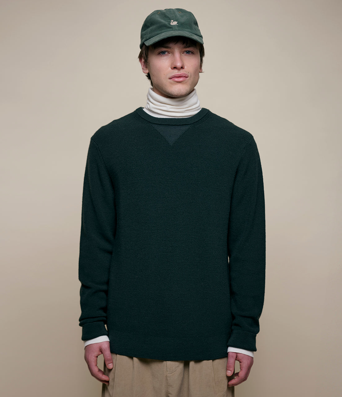 Person wearing a dark green sweater, beige pants, and a matching cap on a beige background