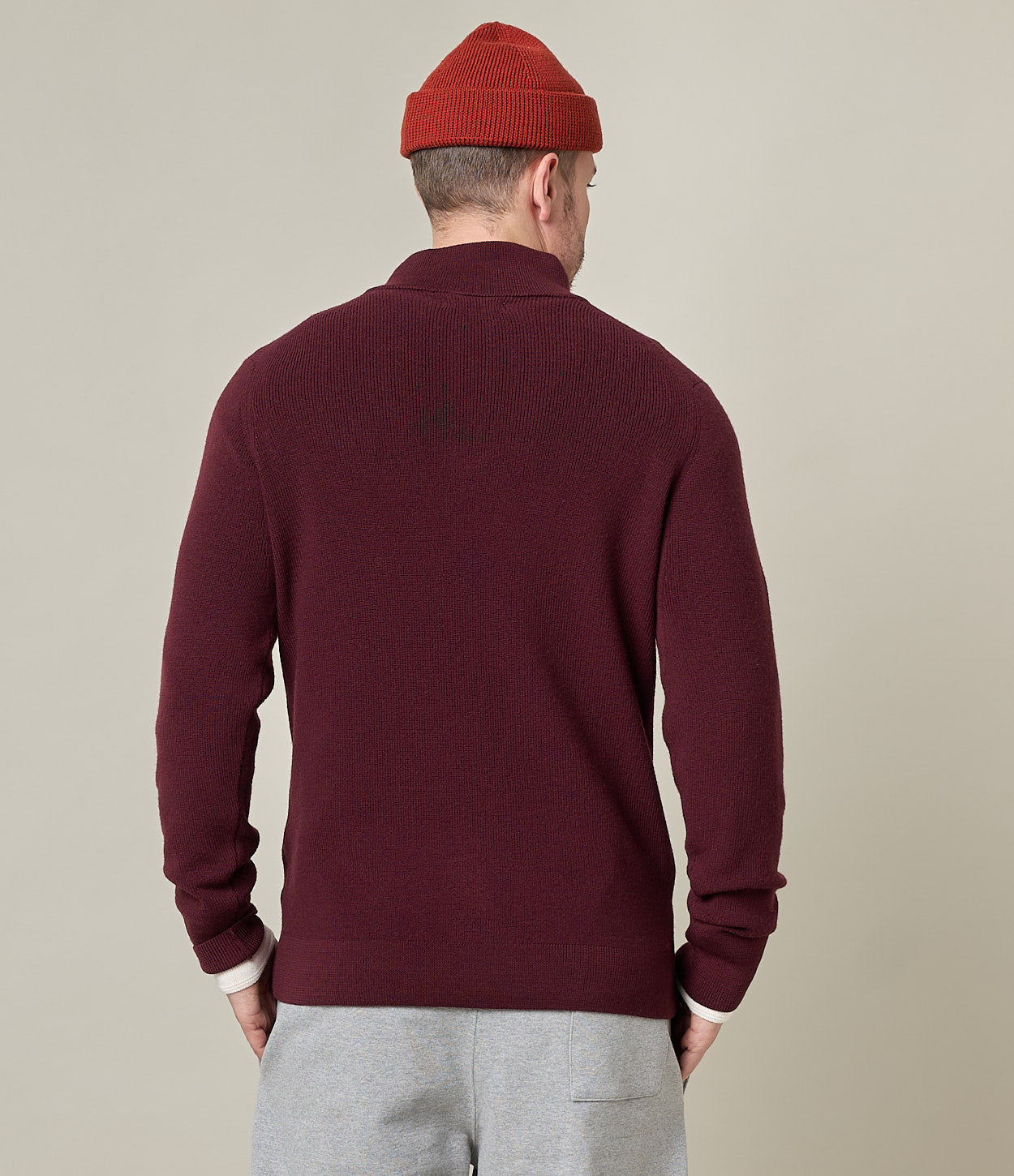 Man wearing a burgundy sweater and red beanie from behind on a beige background