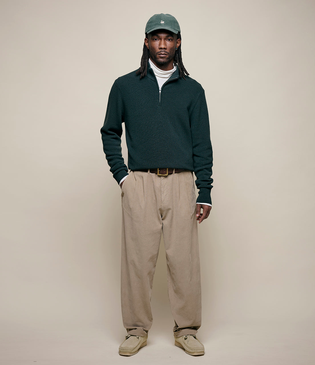 Man wearing a dark green sweater, beige pants, and a cap against a plain background