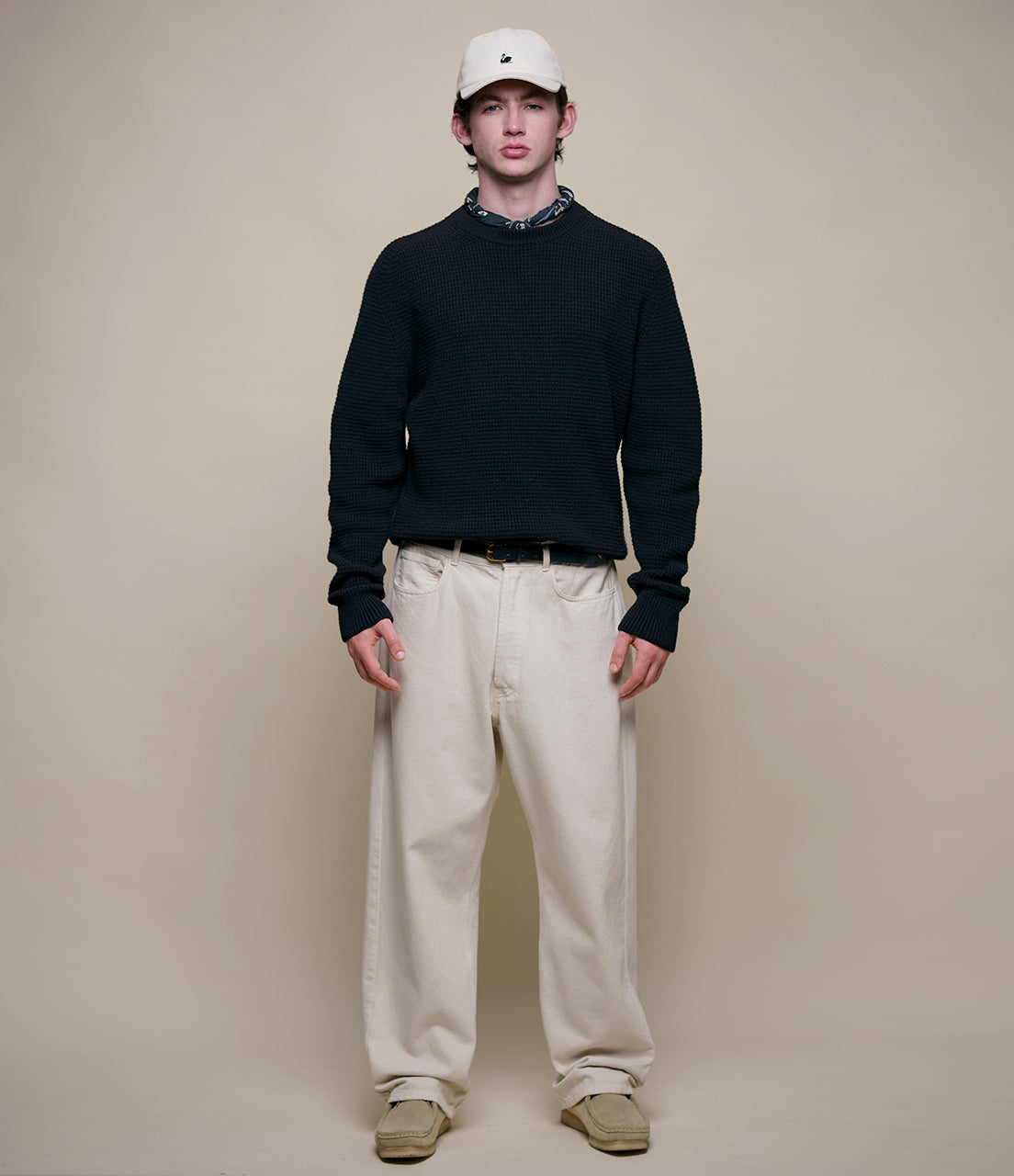 Man wearing a black sweater, beige pants, and a white cap against a plain background