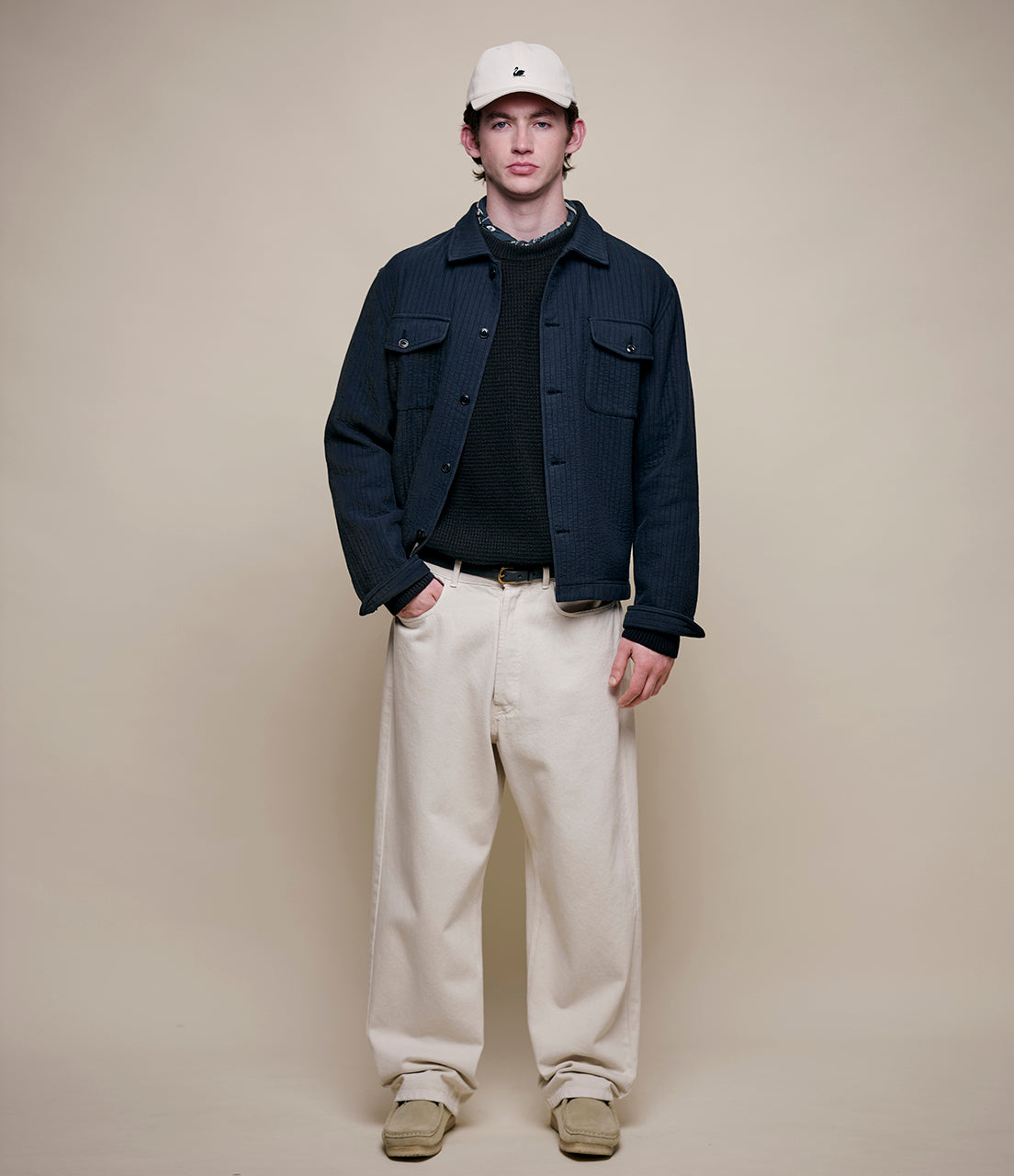 Man wearing a navy jacket, black shirt, beige pants, and white cap against a beige background