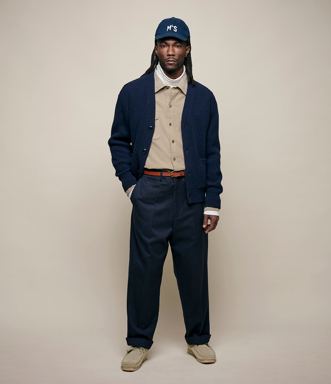 Man wearing a navy cardigan, beige shirt, dark pants, and cap on a plain background