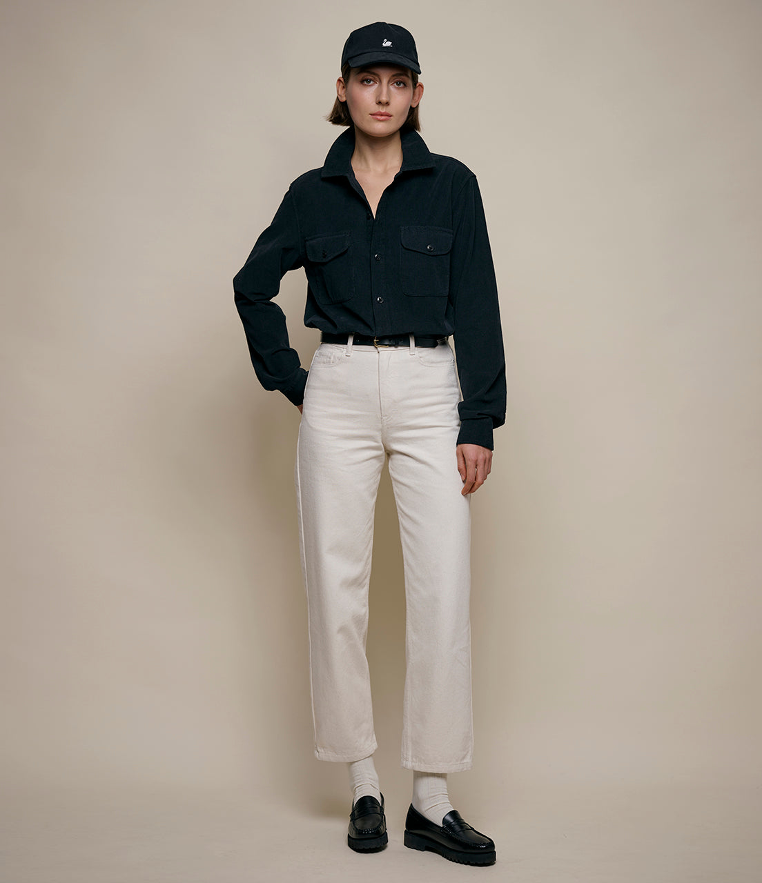 Woman wearing a black corduroy shirt, white pants, and black shoes against a beige background