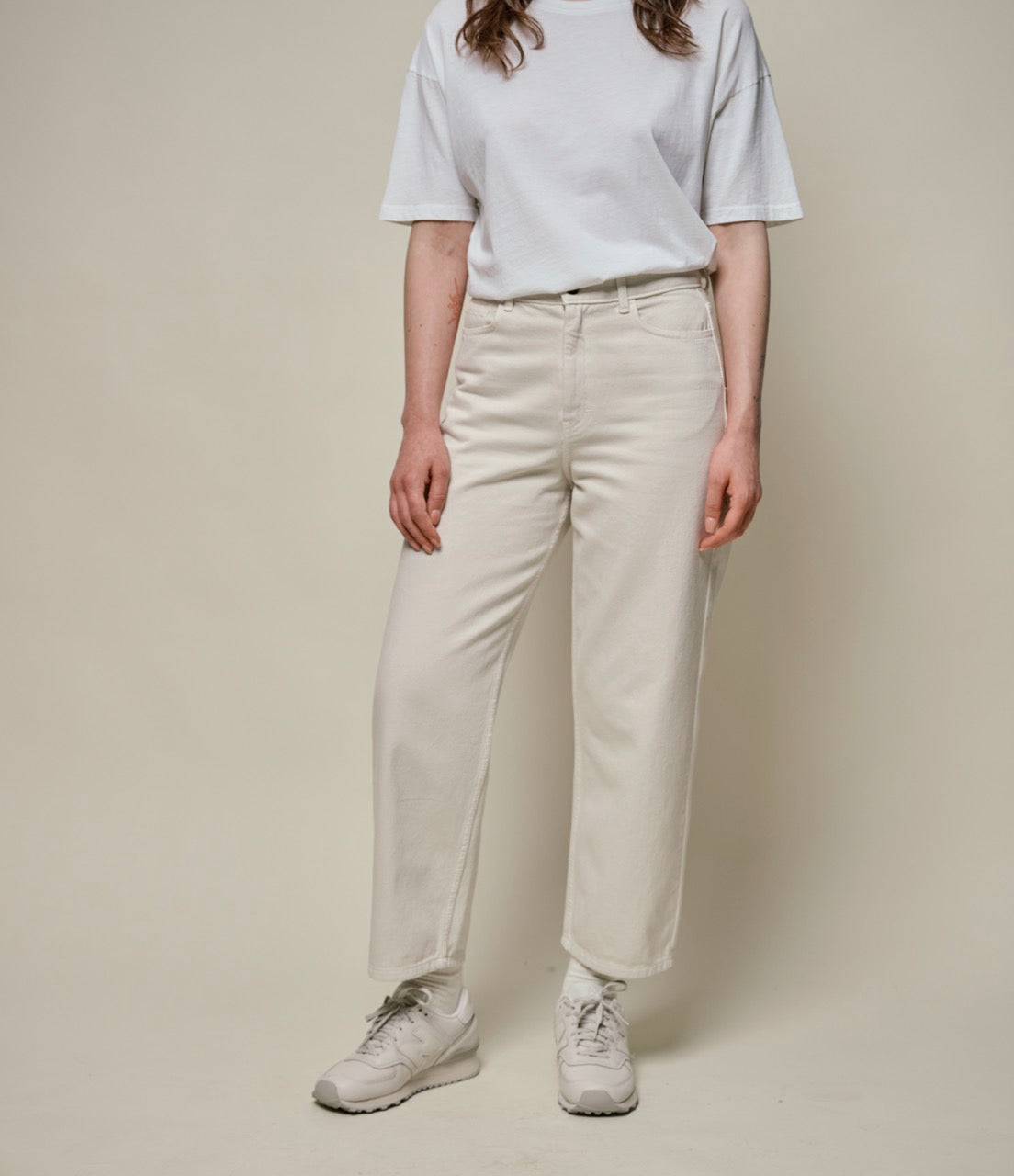 A person standing against a beige background, wearing white ankle-length 5-pocket-pants and a white t-shirt, with no visible shoes.