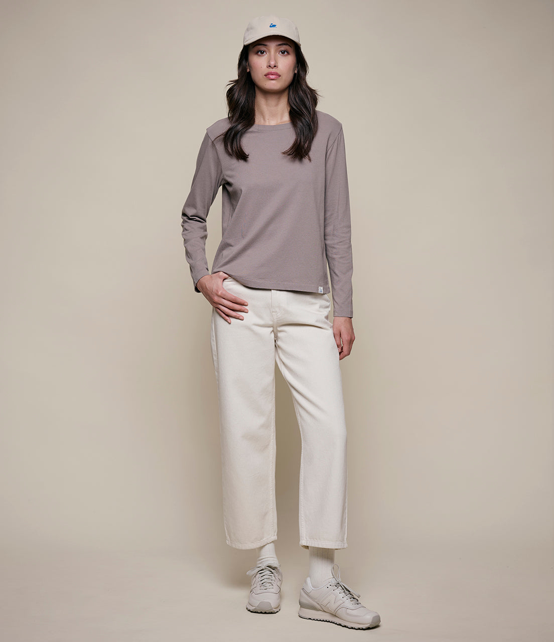 Woman wearing a gray long-sleeve shirt, white pants, and a cap on a beige background