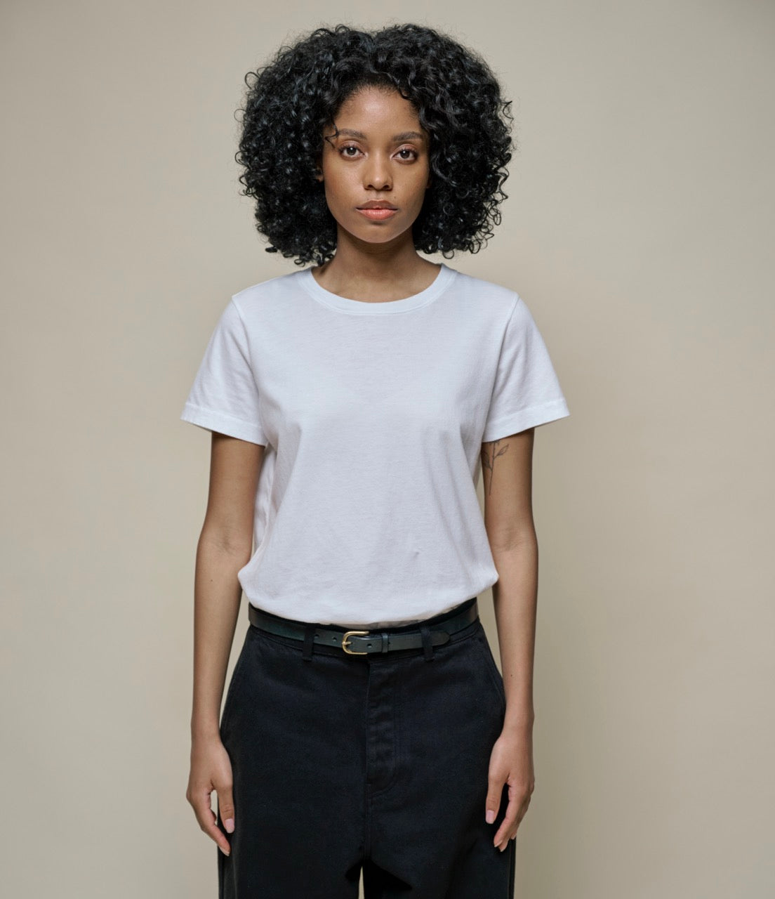 A woman standing against a plain background, wearing a white crew-neck T-shirt and black pants.