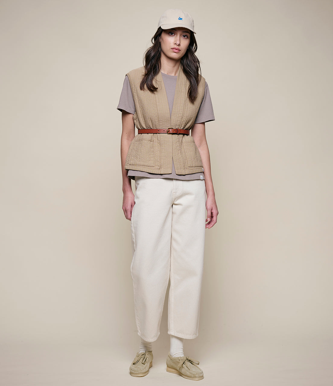 Woman wearing a beige vest, white pants, and a cap on a plain background