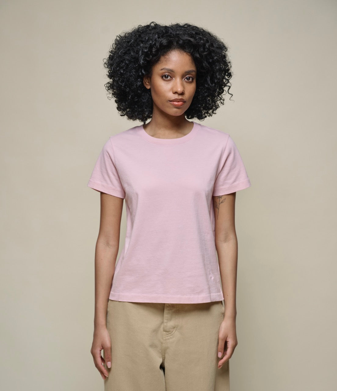 A woman standing against a beige background, wearing a pink, short-sleeved, round-neck T-shirt with no visible brand logo.