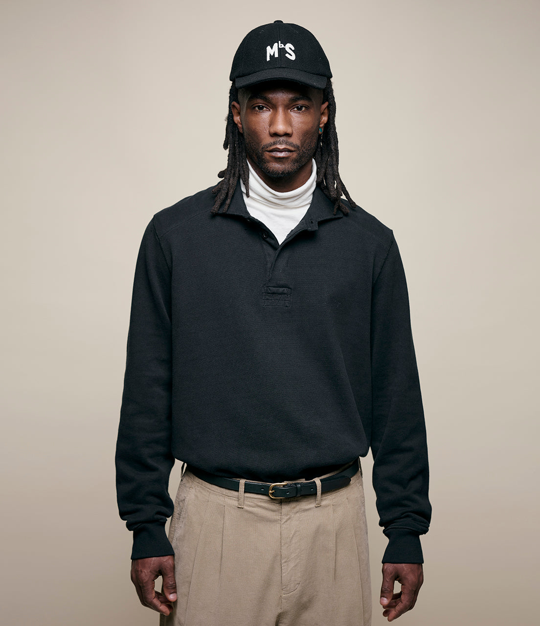 Man wearing a black rugby shirt, beige pants, and a black cap with 'MbS' logo on a beige background
